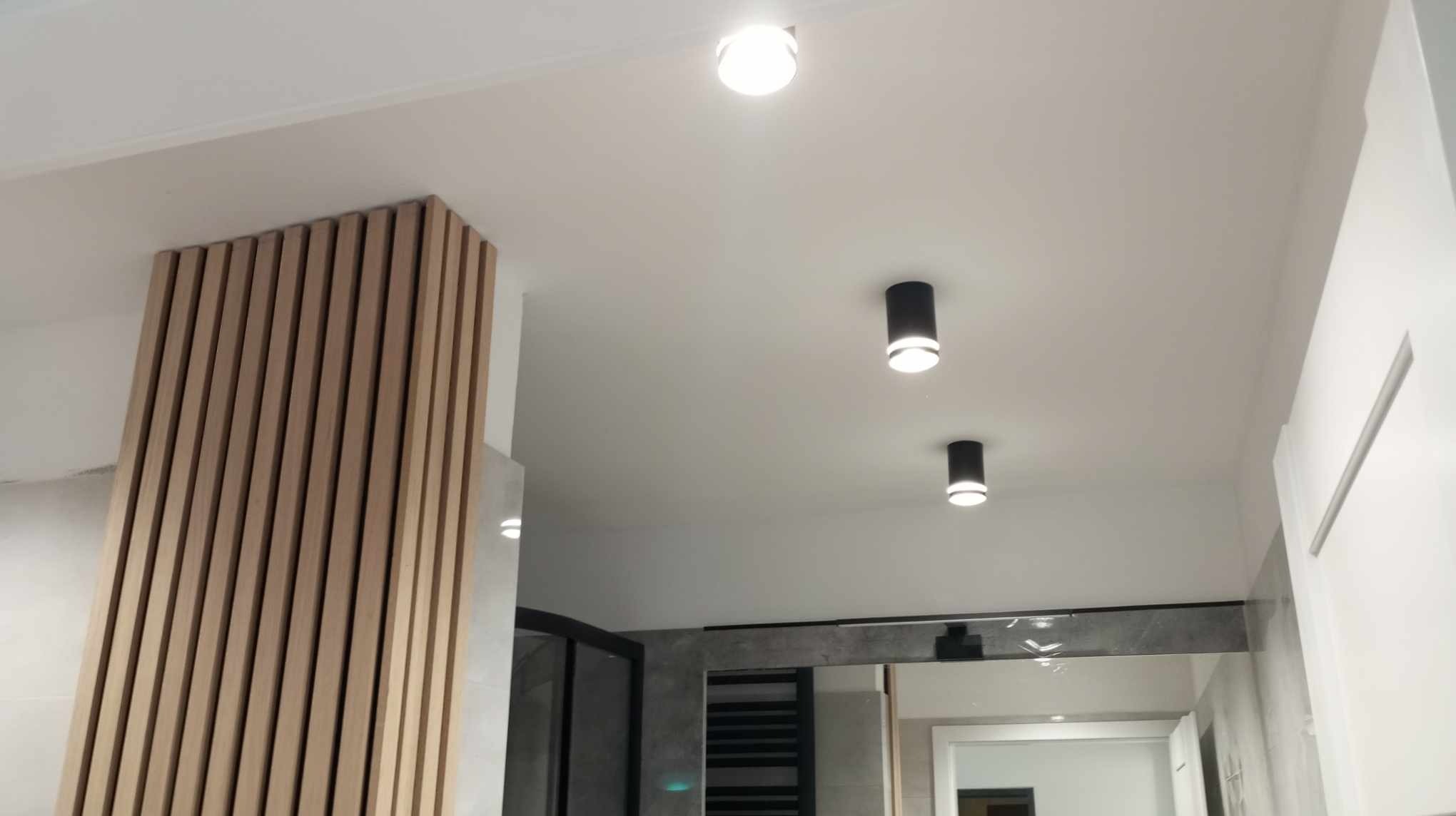 Nowoczesna łazienka z drewnianą ścianką działową z lameli, sufit z trzema punktowymi lampami, widoczne elementy kabiny prysznicowej i grzejnika.