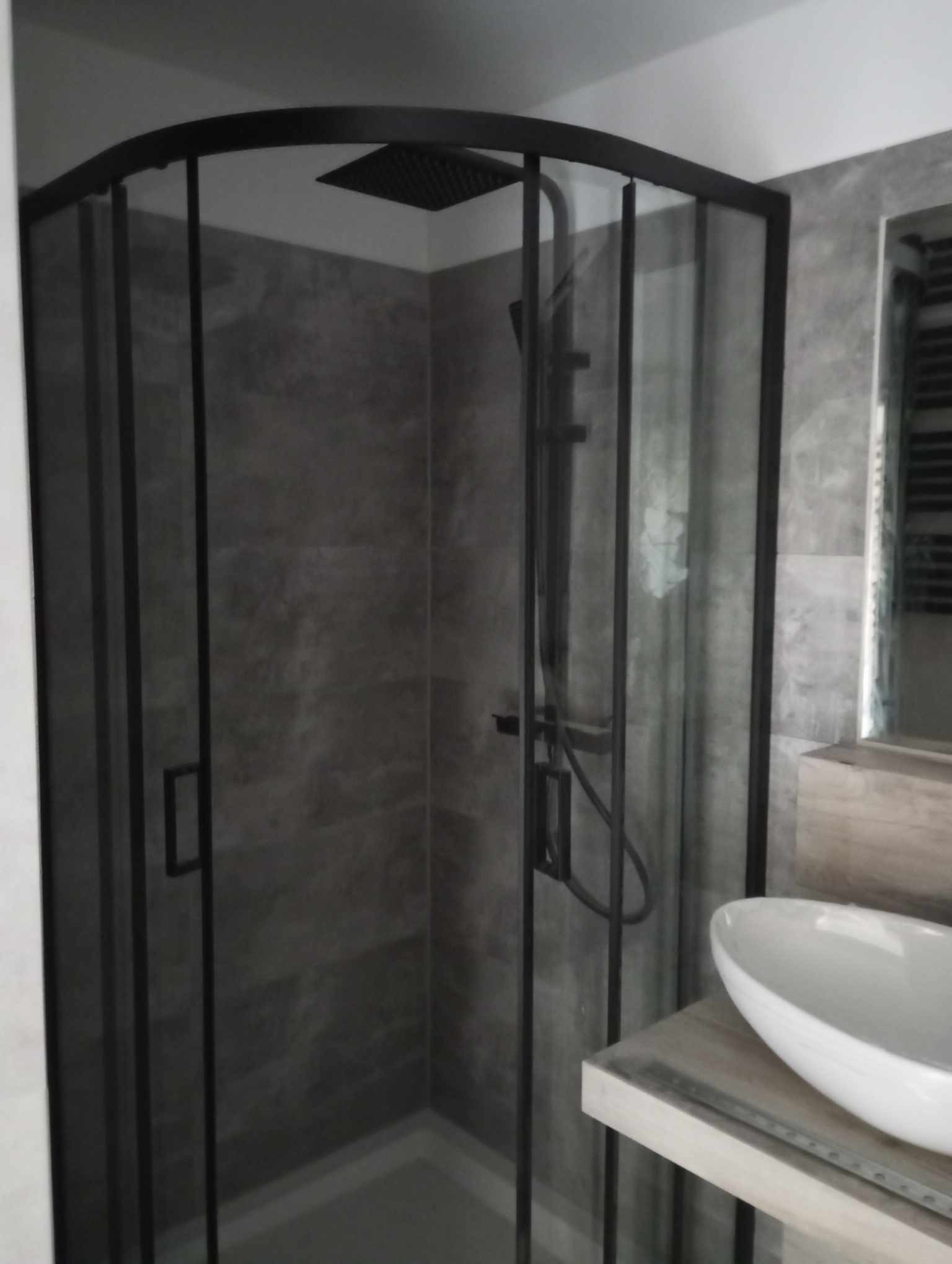 Nowoczesna kabina prysznicowa z czarnymi profilami i deszczownicą, szare płytki imitujące beton na ścianach, biała umywalka nablatowa na drewnianym blacie, minimalistyczny styl łazienki.