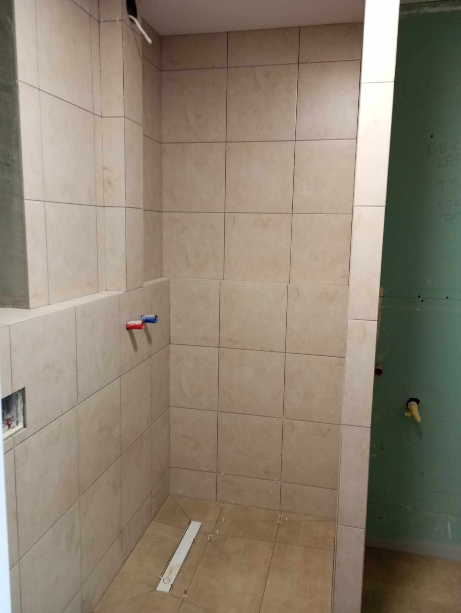 Niedokończona łazienka z beżowymi płytkami, widoczny odpływ liniowy w podłodze i wystające rury instalacji wodnej na ścianie z płyt gipsowo-kartonowych z napisem.