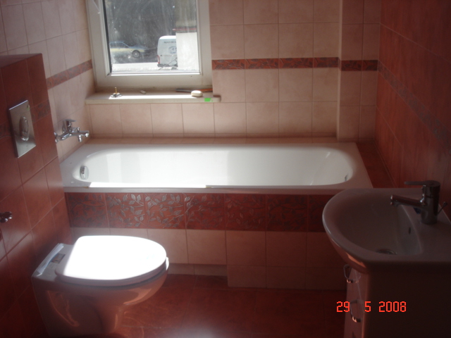 Ukończona łazienka w odcieniach brązu i beżu z wanną, umywalką i podwieszanym WC, wykończona płytkami z dekoracyjnym motywem roślinnym na obudowie wanny.