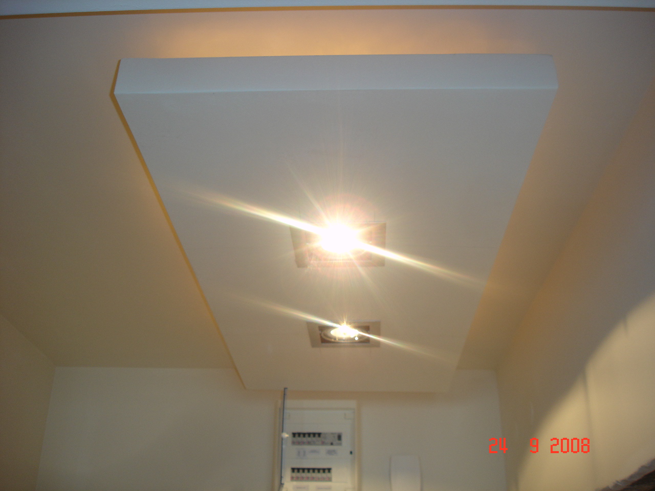 Wnętrze z podwieszanym sufitem z dwoma punktami świetlnymi, widoczna instalacja elektryczna na ścianie, ściany w odcieniach bieli.