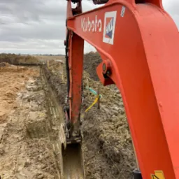 Pomarańczowa koparka Kubota w trakcie wykonywania wykopu w grząskiej, błotnistej glebie; widoczne drewniane paliki i napięty sznurek wyznaczające linię wykopu, pochmurne niebo w tle.
