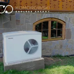 Biała pompa ciepła Stiebel Eltron zamontowana na betonowym fundamencie przy kamiennej ścianie budynku z drewnianym oknem; widoczne logo firmy Komorowski Endless Energy na elewacji.