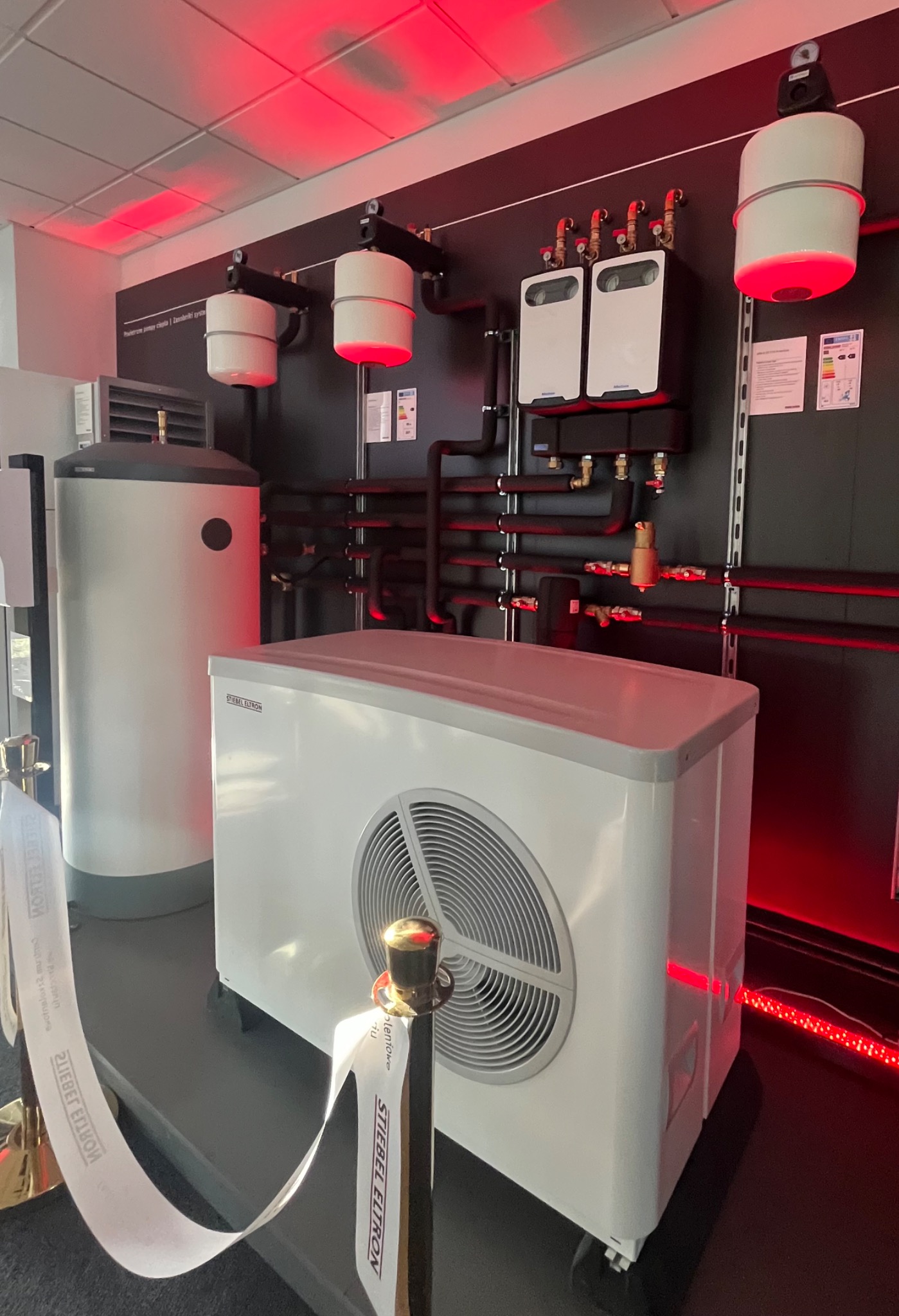 Wystawa systemu grzewczego Stiebel Eltron: pompa ciepła obok zasobnika wody, czarne rury instalacji i białe moduły na ścianie, oświetlone czerwonym światłem.