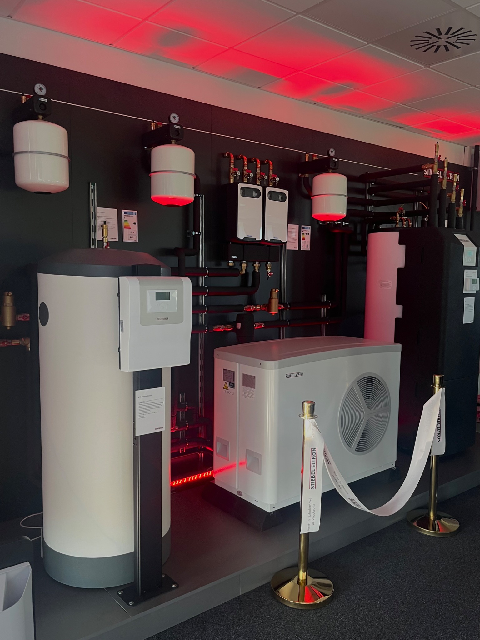 Ekspozycja urządzeń grzewczych Stiebel Eltron w showroomie: pompa ciepła, zbiornik buforowy i zasobnik ciepłej wody, połączone rurami w systemie, podświetlone czerwonym światłem