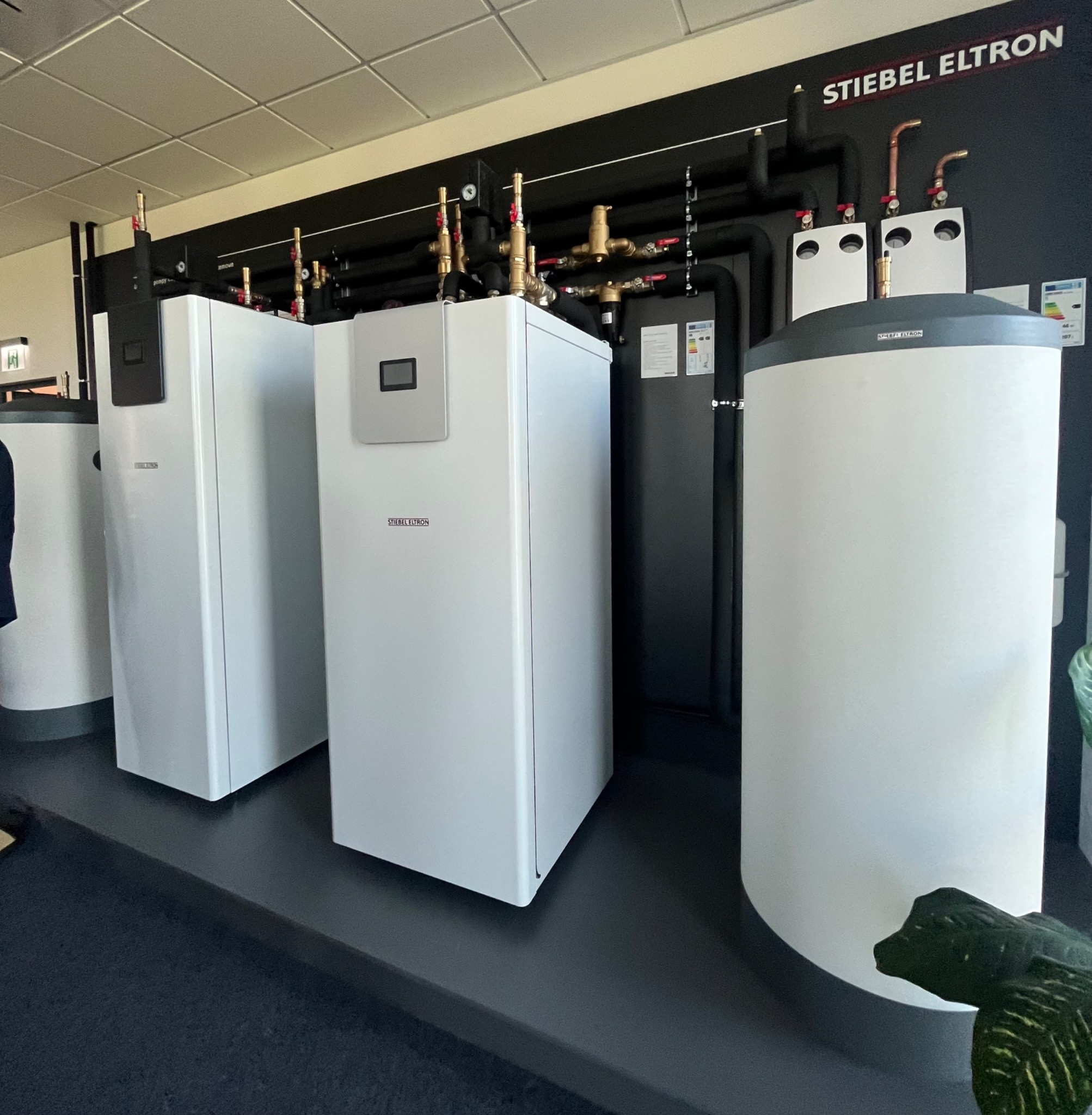 Ekspozycja kilku białych pomp ciepła marki Stiebel Eltron z czarnymi rurami i zaworami, ustawionych na czarnym podeście, z fragmentem rośliny w prawym dolnym rogu.