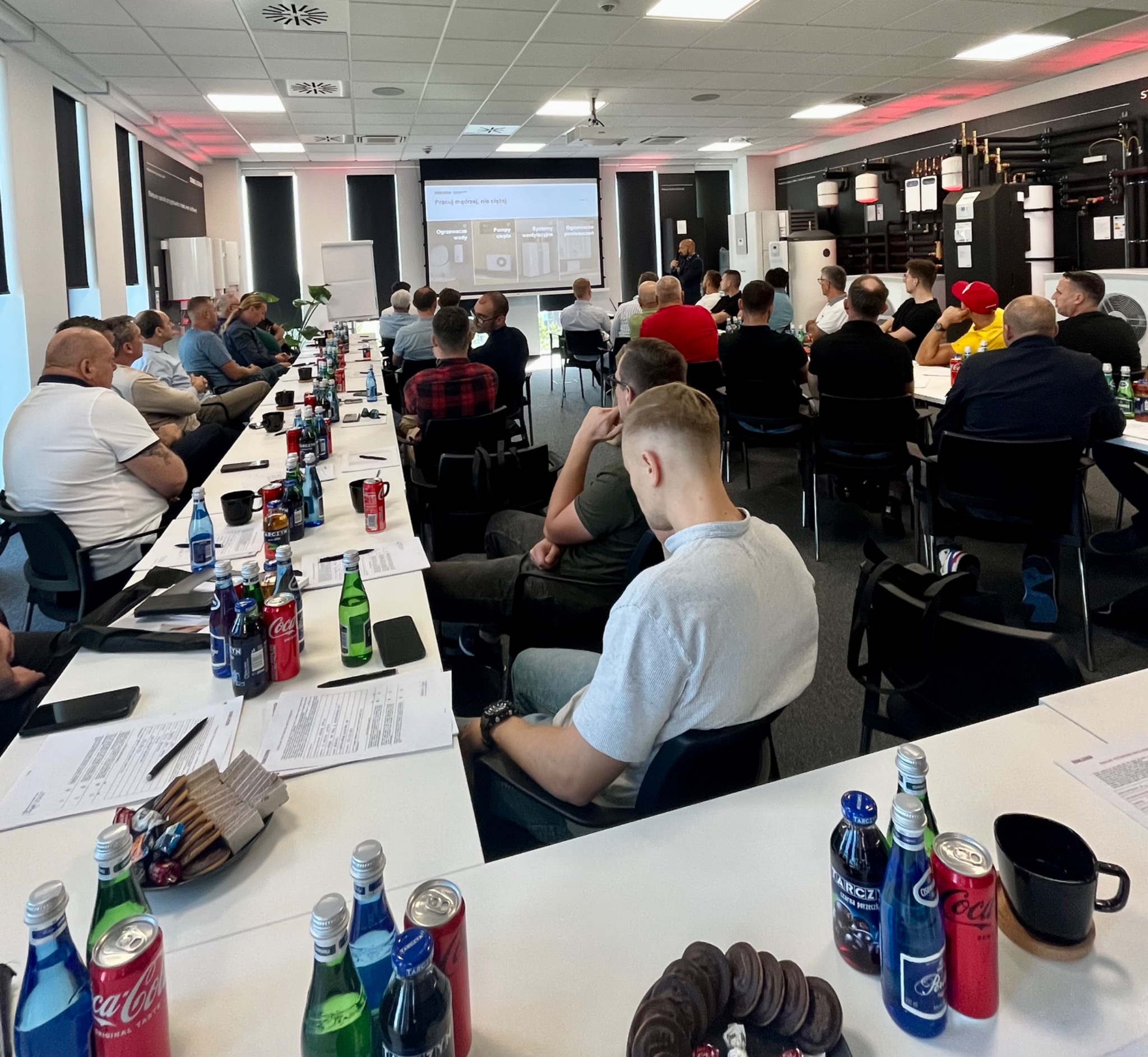 Sala konferencyjna z uczestnikami szkolenia, na ścianie ekspozycja urządzeń grzewczych, na stołach napoje i przekąski, na ekranie prezentacja z grafikami urządzeń grzewczych.