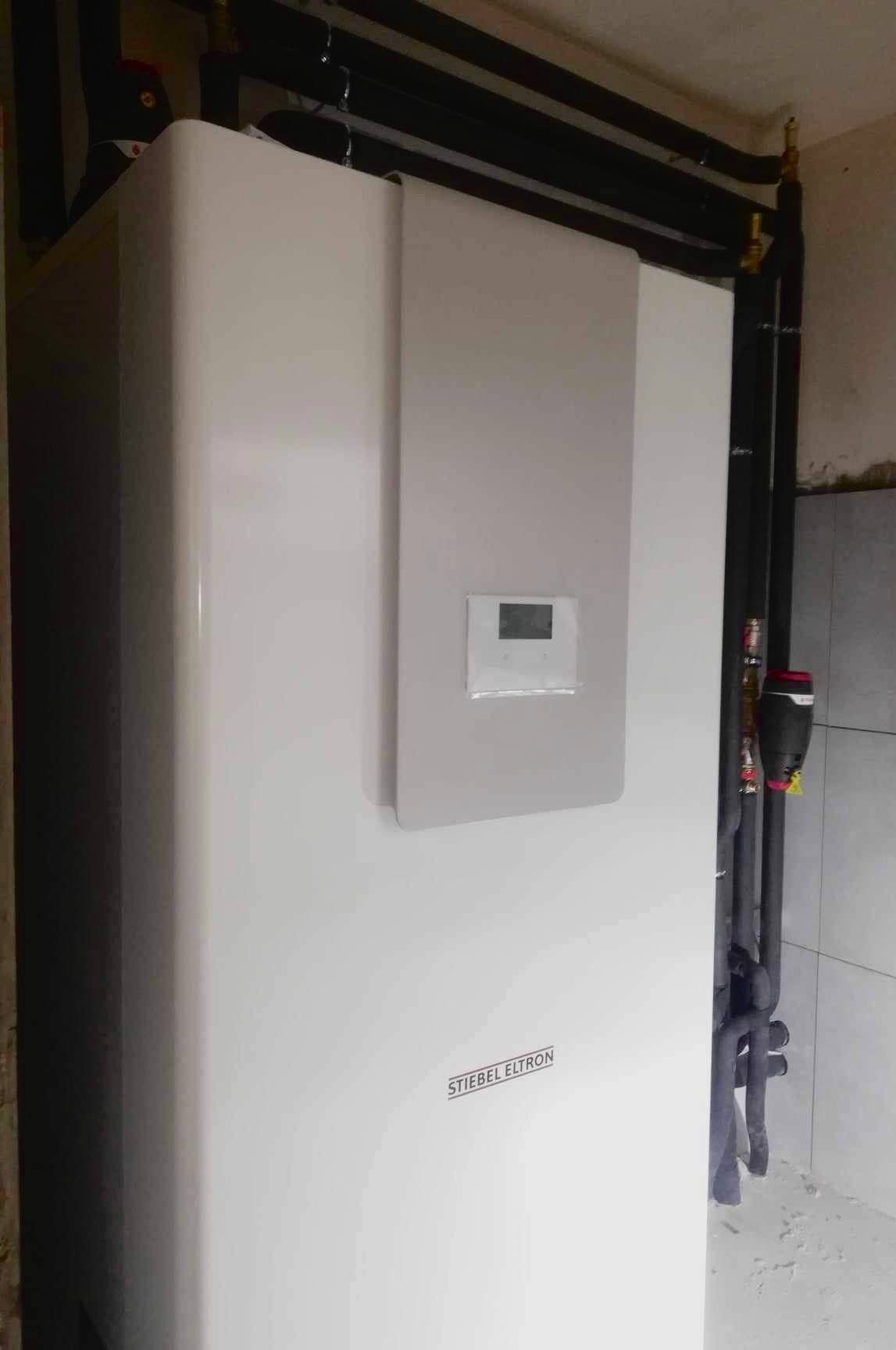 Biała pompa ciepła Stiebel Eltron z czarnymi rurami instalacyjnymi w tle, widoczne elementy instalacji grzewczej.