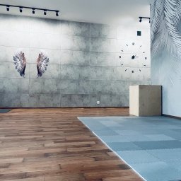 MadaFit Magdalena Doniecka - Jasne, przestronne wnętrze sali do ćwiczeń z drewnianą podłogą, szarą ścianą z betonowych paneli z rysunkiem skrzydeł, dużym zegarem i podłogą z szarych puzzli piankowych. W rogu drewniany sześcian.