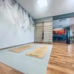 Jasna sala do ćwiczeń z drabinką gimnastyczną, dwoma matami do ćwiczeń leżącymi na podłodze z piankowych puzzli, ściana z motywem liści palmowych, sprzęt do ćwiczeń w tle.