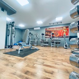 MadaFit Magdalena Doniecka - Wnętrze siłowni z różnorodnym sprzętem do ćwiczeń siłowych, w tle plakat z wizerunkiem trenerki Magdaleny Krajkowskiej, na pierwszym planie regał z chromowanymi hantlami.