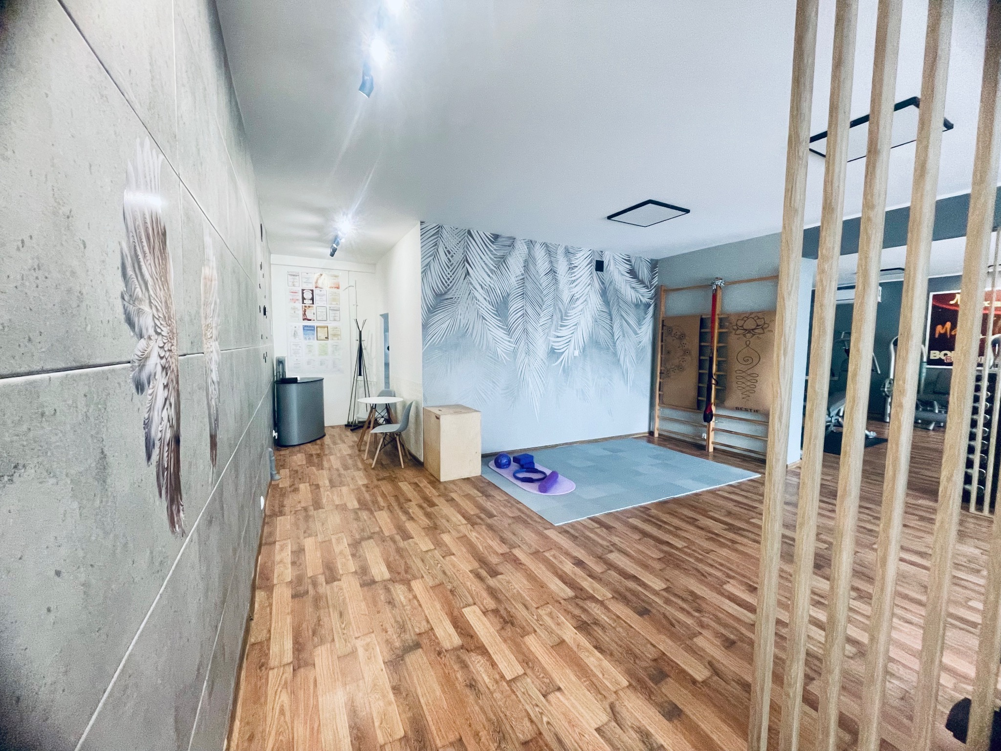 Jasne, przestronne studio z drewnianą podłogą, drabinkami gimnastycznymi, ścianą z motywem liści palmowych, matą do ćwiczeń, bloczkami i paskiem do jogi oraz ścianą z grafiką skrzydeł.
