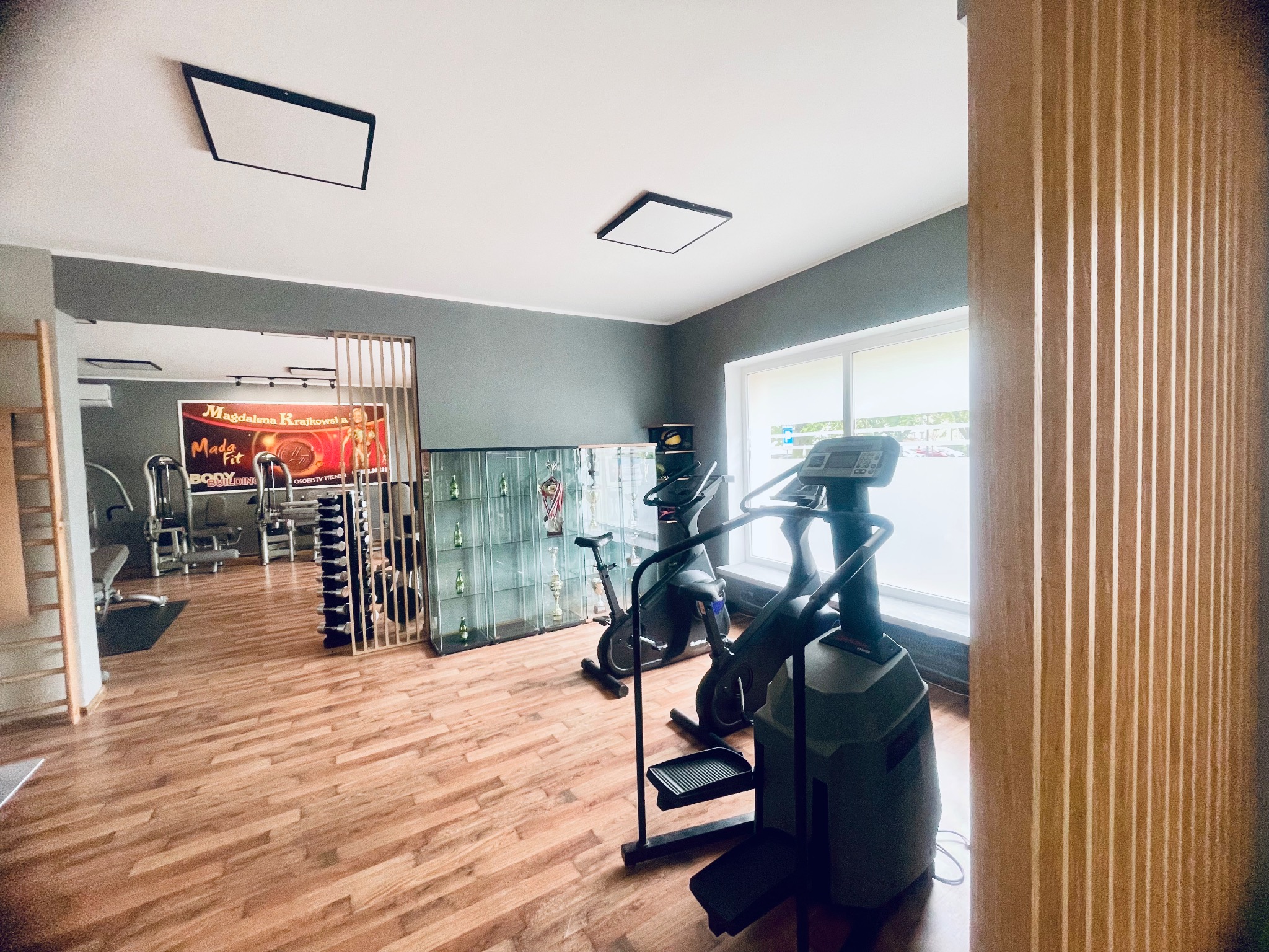Wnętrze siłowni z parkietem, widoczne maszyny do ćwiczeń, rower stacjonarny, schody treningowe oraz regał z trofeami i plakat z wizerunkiem kobiety.