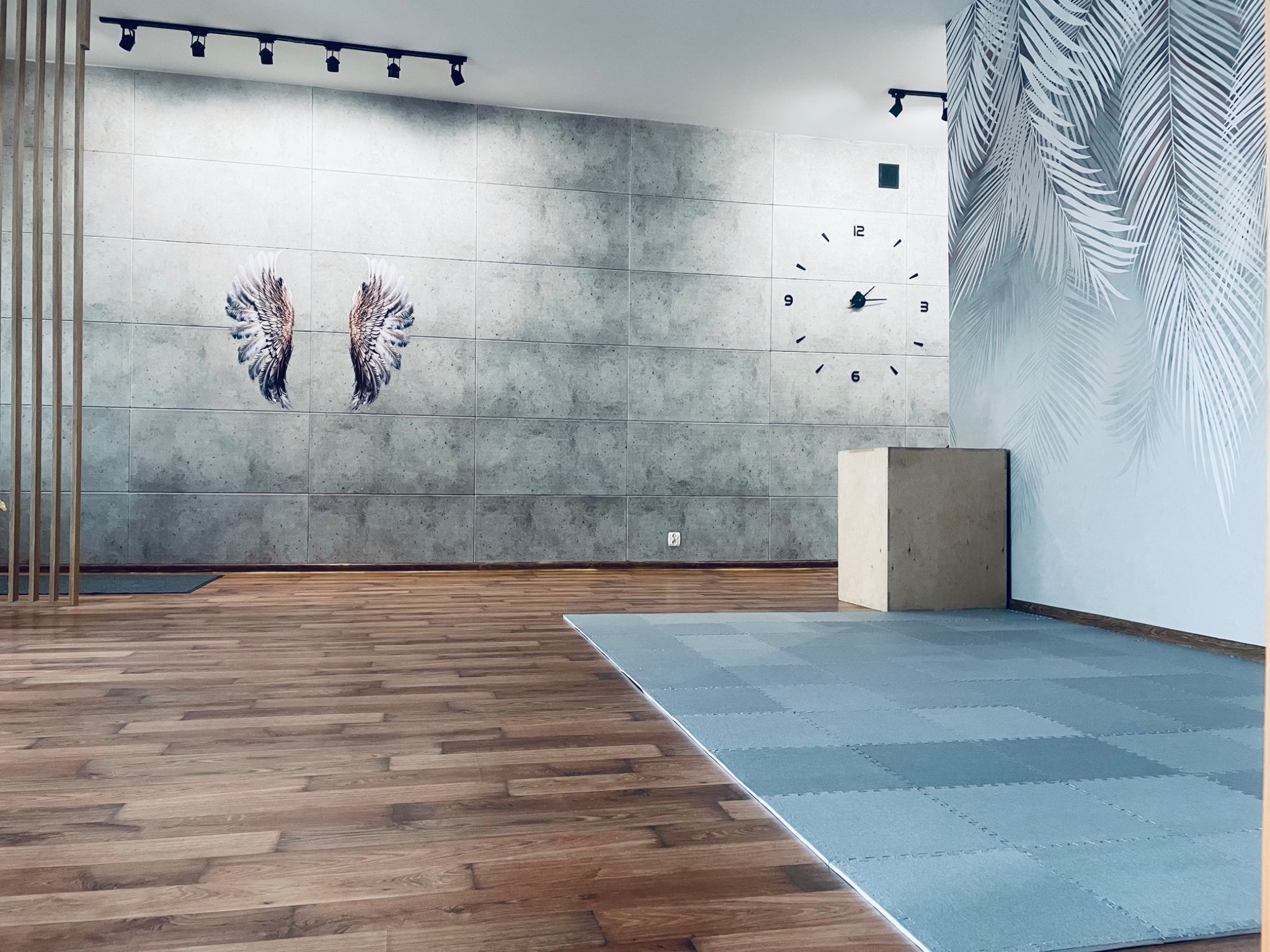 Jasne, przestronne wnętrze sali do ćwiczeń z drewnianą podłogą, szarą ścianą z betonowych paneli z rysunkiem skrzydeł, dużym zegarem i podłogą z szarych puzzli piankowych. W rogu drewniany sześcian.