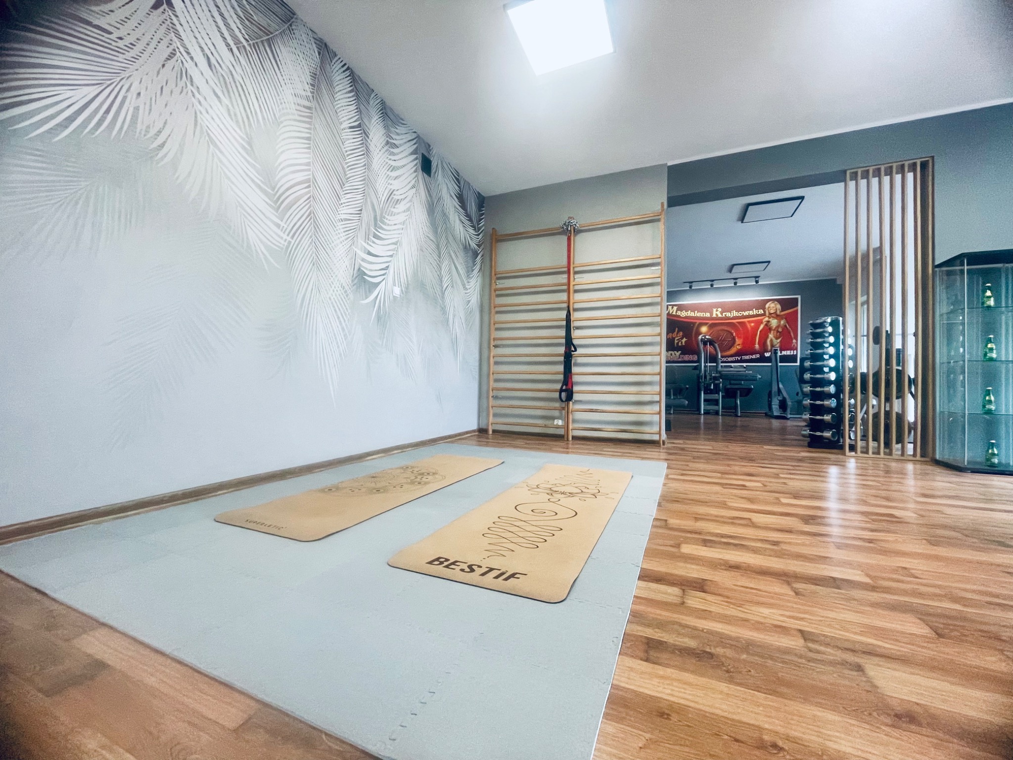 Jasna sala do ćwiczeń z drabinką gimnastyczną, dwoma matami do ćwiczeń leżącymi na podłodze z piankowych puzzli, ściana z motywem liści palmowych, sprzęt do ćwiczeń w tle.