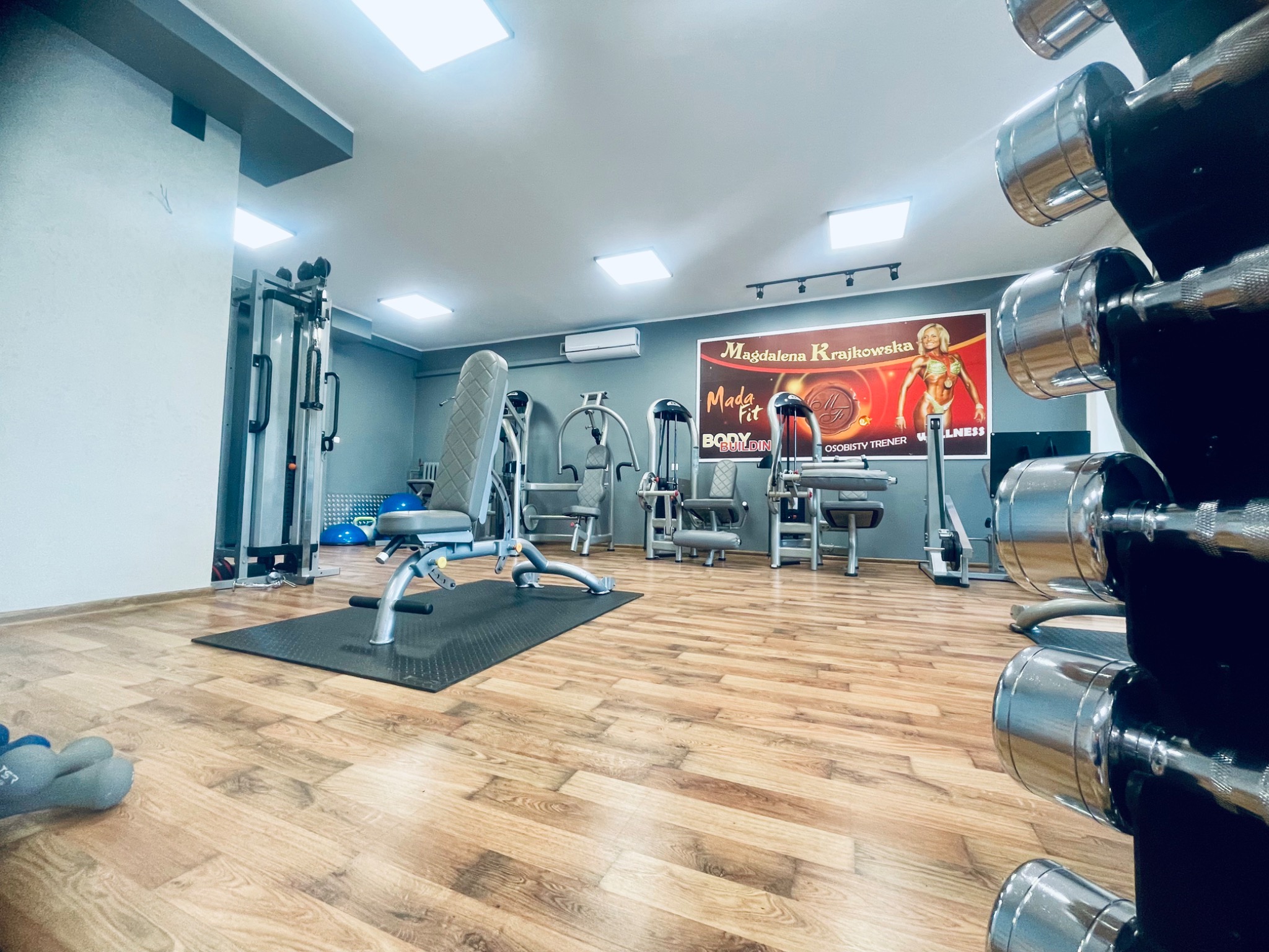 Wnętrze siłowni z różnorodnym sprzętem do ćwiczeń siłowych, w tle plakat z wizerunkiem trenerki Magdaleny Krajkowskiej, na pierwszym planie regał z chromowanymi hantlami.