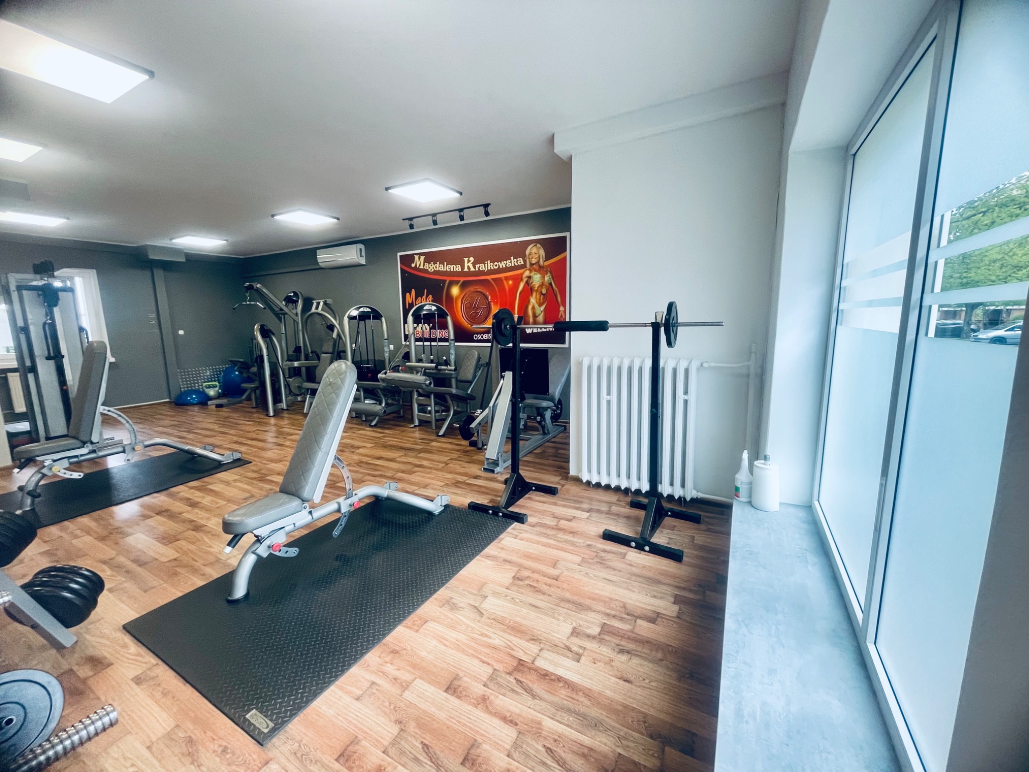 Wnętrze siłowni z różnorodnym sprzętem do ćwiczeń siłowych, w tle plakat z wizerunkiem kobiety i logo. Widoczny sztanga na stojaku przy oknie.