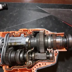 Wnętrze przekładni pomarańczowej obudowy urządzenia budowlanego, widoczne metalowe zębatki i elementy mechaniczne pokryte smarem.