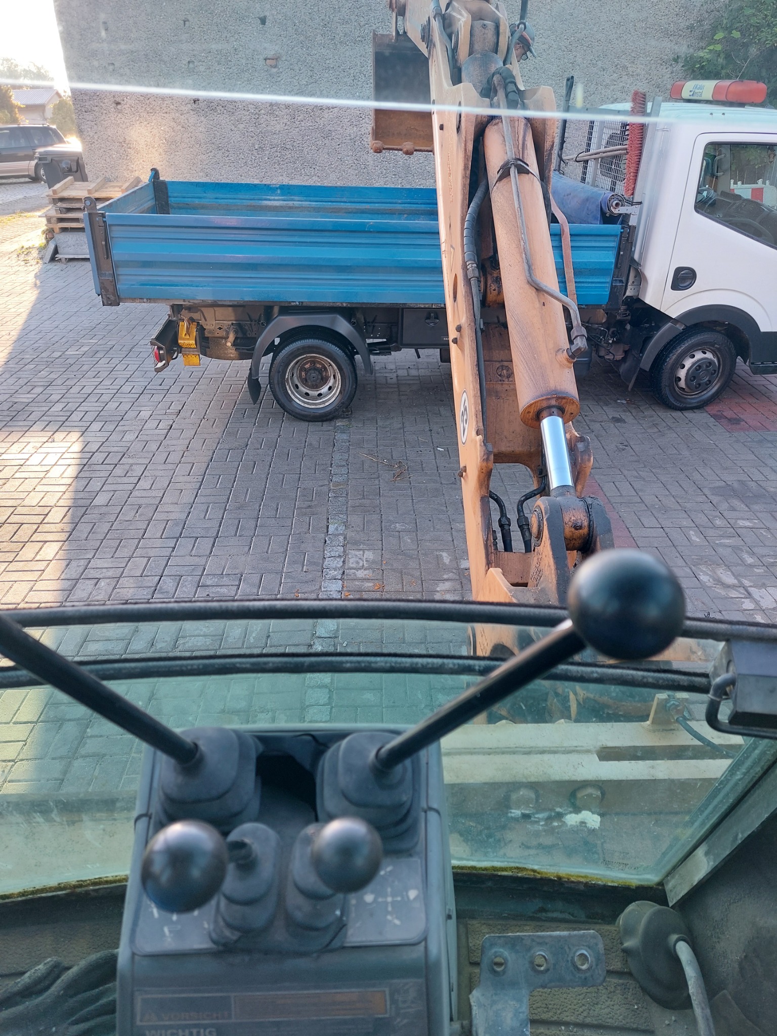 Widok z kabiny koparki na pomarańczowe ramię i białą ciężarówkę z niebieską skrzynią ładunkową, zaparkowaną na kostce brukowej.