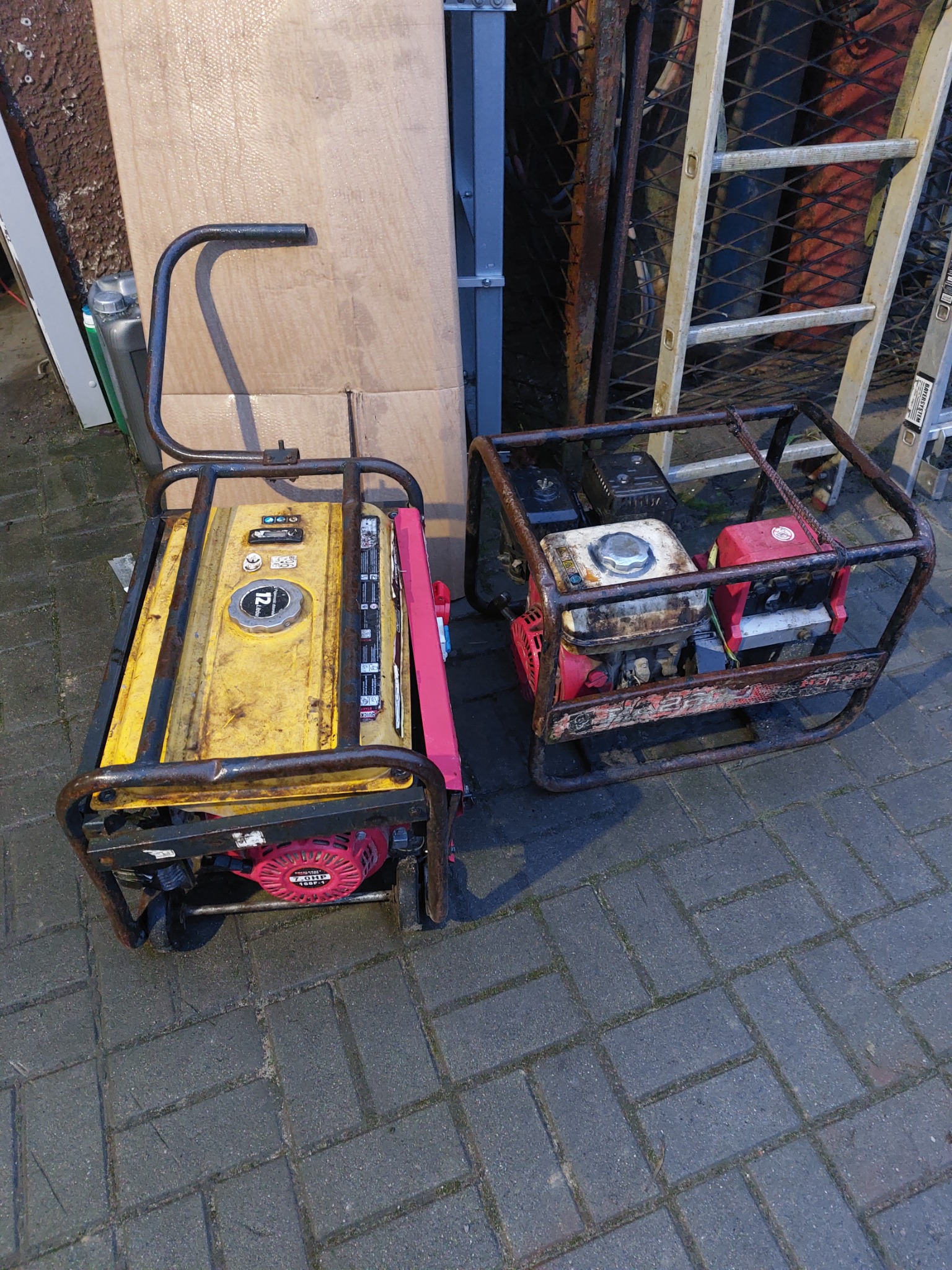 Dwa używane agregaty prądotwórcze na brukowanej powierzchni, widoczne ślady użytkowania i korozji, w tle drabina i płyta OSB.