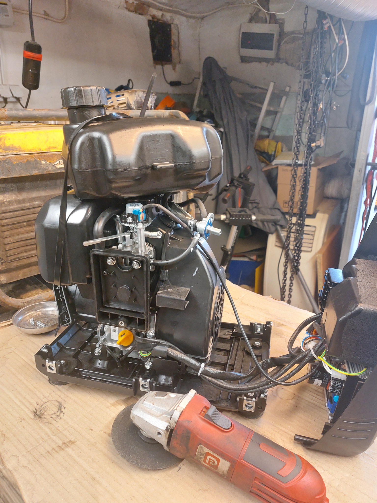 Czarny silnik spalinowy z odkręconą obudową i szlifierką kątową obok, na drewnianym stole warsztatowym w zagraconym garażu.