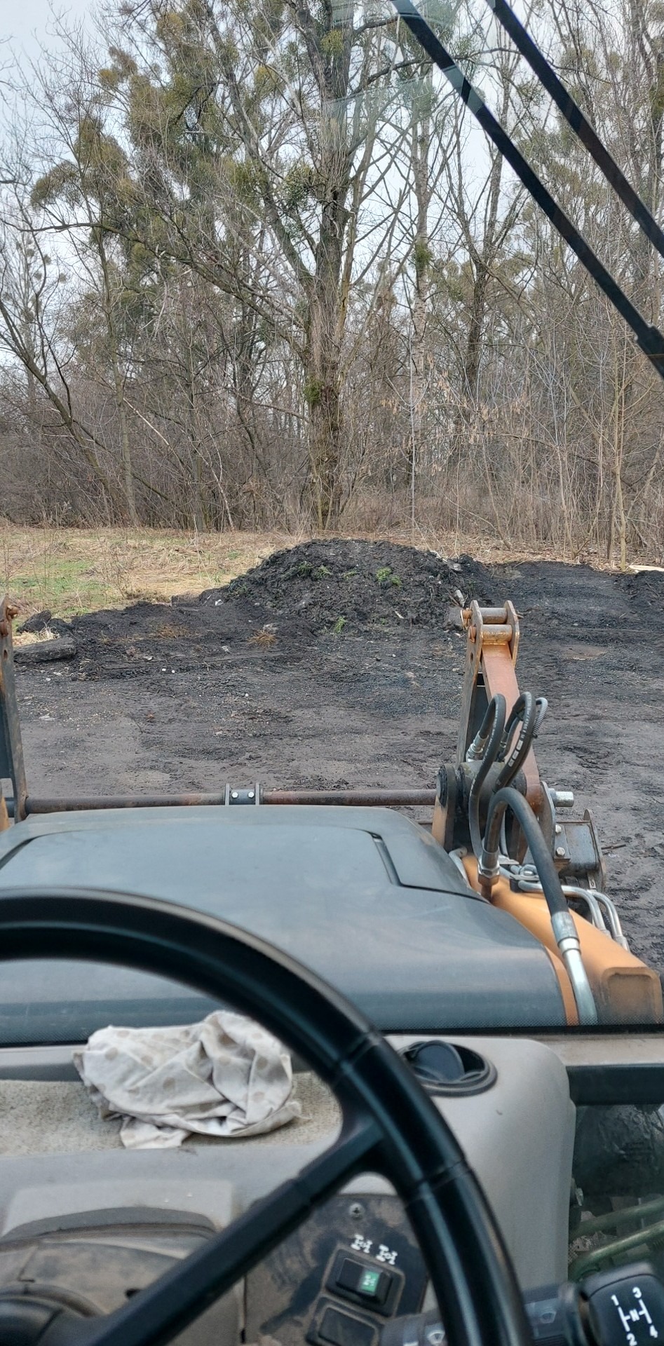 Widok z kabiny koparko-ładowarki na pryzmę ziemi i drzewa w tle, z widoczną deską rozdzielczą i kierownicą.