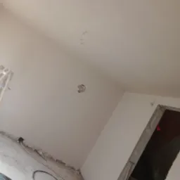 Wnętrze w trakcie remontu; ściany i sufit po nałożeniu gładzi, widoczne niedoskonałości i otwór w ścianie, kabel wystający z sufitu, podłoga z resztkami gruzu i kablami.