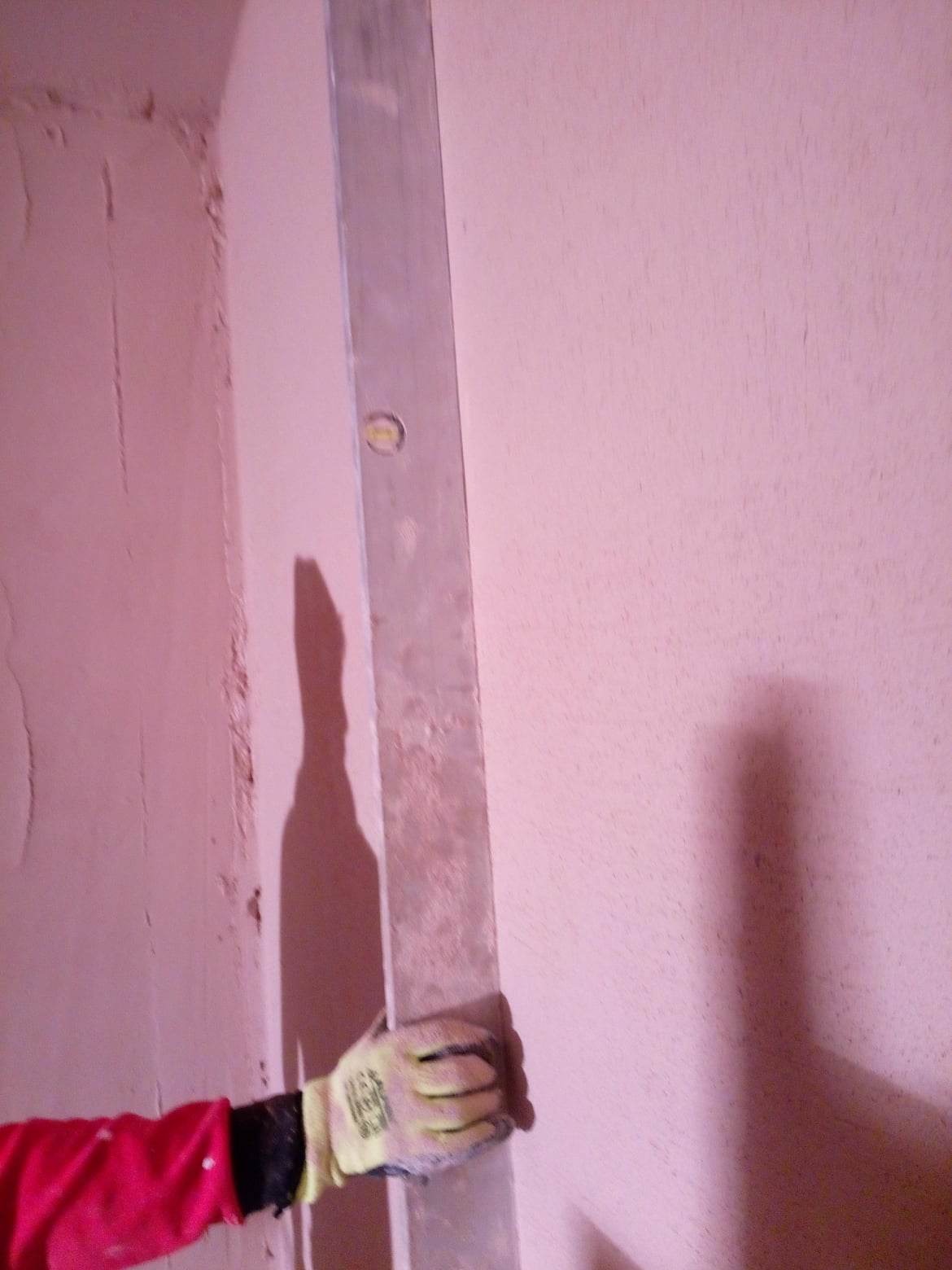 Pracownik w rękawicach wyrównuje ścianę za pomocą długiej poziomicy, widoczna faktura ściany przed wygładzeniem.