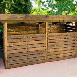 Drewniana wiata śmietnikowa na cztery kubły, z ciemnego drewna, na kostce brukowej, otoczona zielenią.