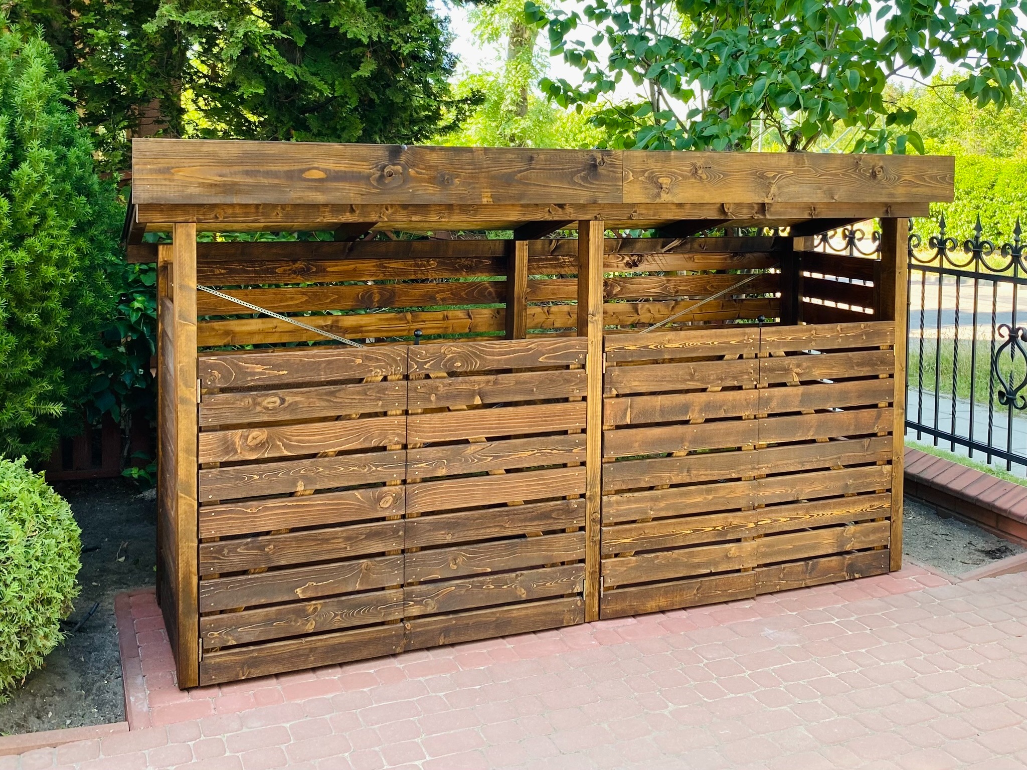 Drewniana wiata śmietnikowa na cztery kubły, z ciemnego drewna, na kostce brukowej, otoczona zielenią.