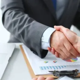 Uścisk dłoni nad dokumentami z wykresami i laptopem, symbolizujący zawarcie umowy lub partnerstwo biznesowe.