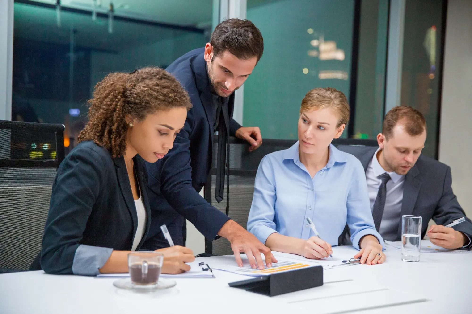 Cztery osoby w biurze analizują dokumenty przy stole. Kobieta z kręconymi włosami pije kawę w szklance podczas notowania. Mężczyzna w garniturze wskazuje na arkusz z danymi.