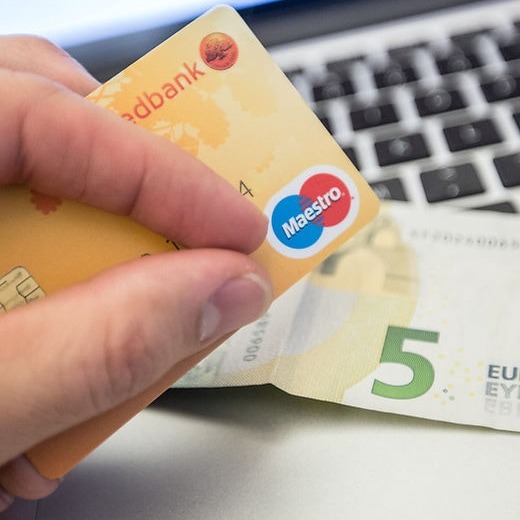 Dłoń trzyma kartę Maestro banku w odcieniach żółci na tle klawiatury laptopa i banknotu 5 euro.