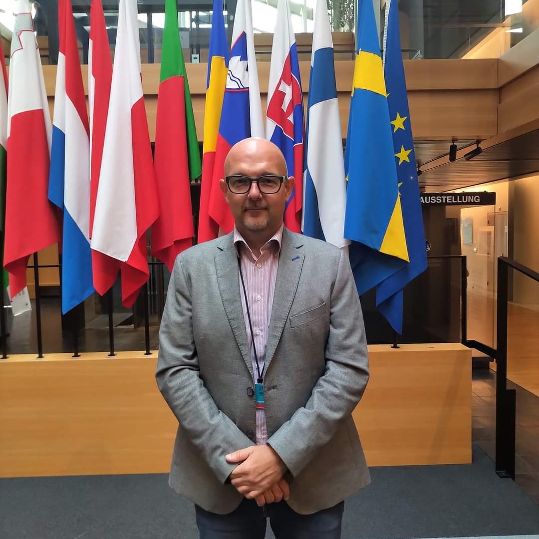 Mężczyzna w szarej marynarce i okularach stoi na tle flag państwowych, z widocznym napisem 'AUSSTELLUNG' w tle.