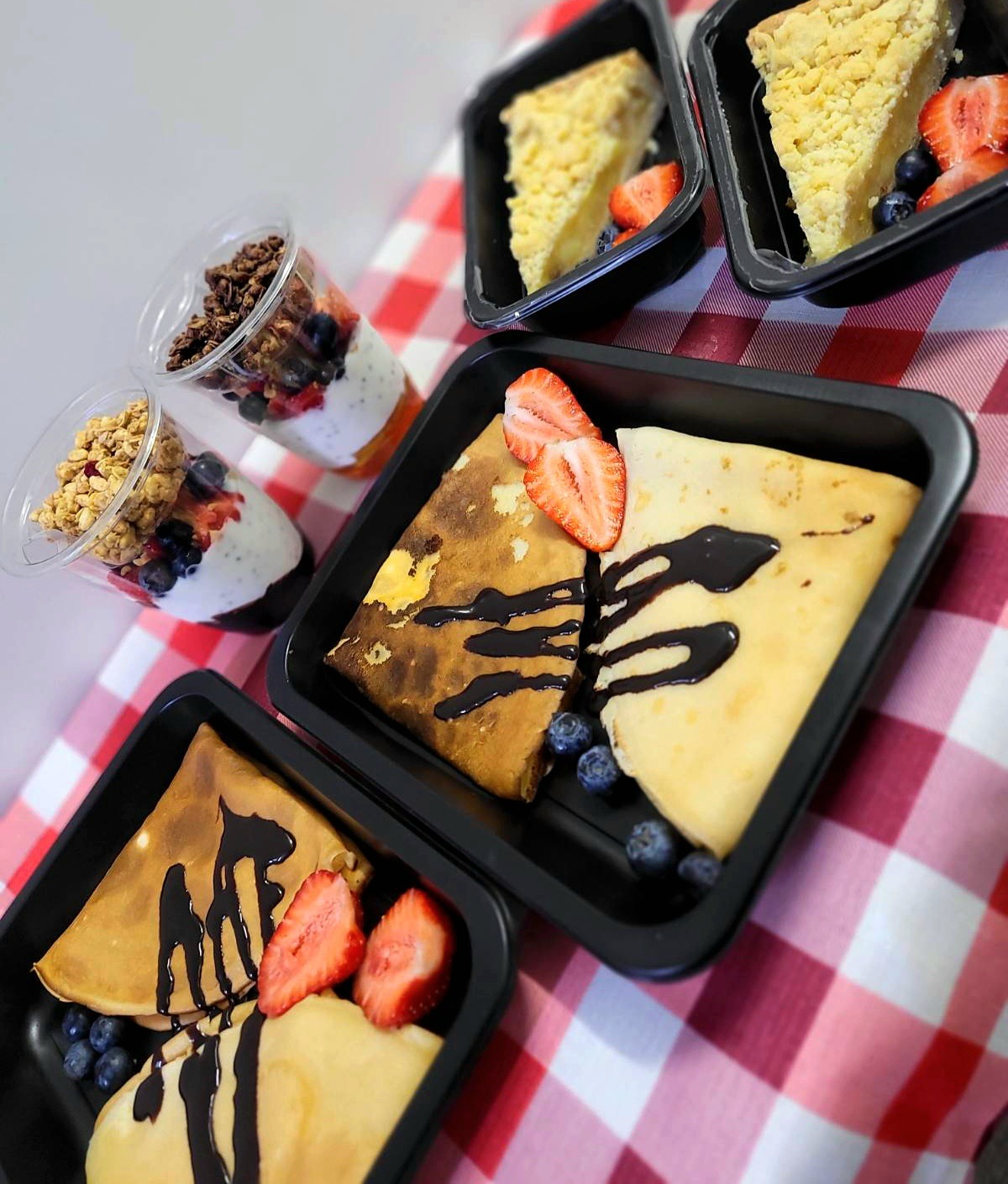 Zestaw posiłków w czarnych pojemnikach: naleśniki z polewą czekoladową i truskawkami, kawałki ciasta z kruszonką oraz desery jogurtowe z granolą i owocami, ułożone na czerwono-białym obrusie w kratę.