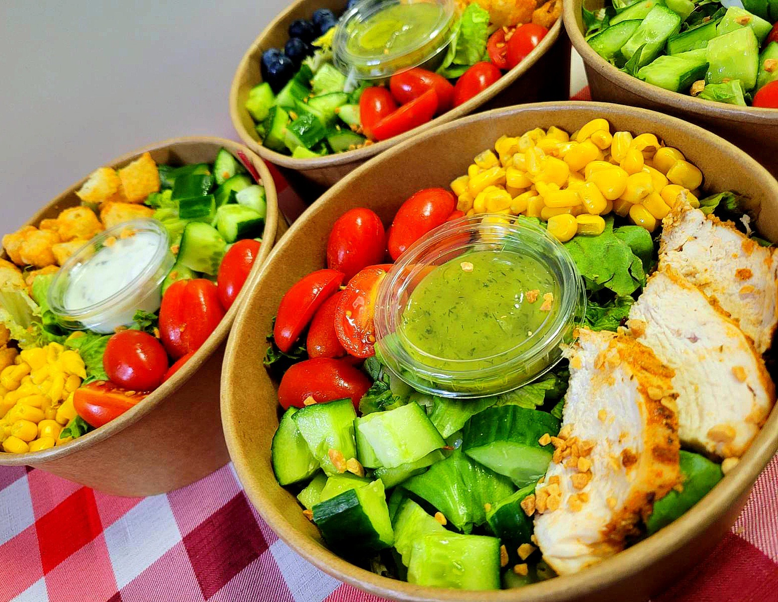 Cztery papierowe miski z różnorodnymi sałatkami: z grillowanym kurczakiem i kukurydzą, z grzankami, z borówkami, każda z osobnym sosem w plastikowym pojemniku, na czerwono-białym obrusie w kratę.