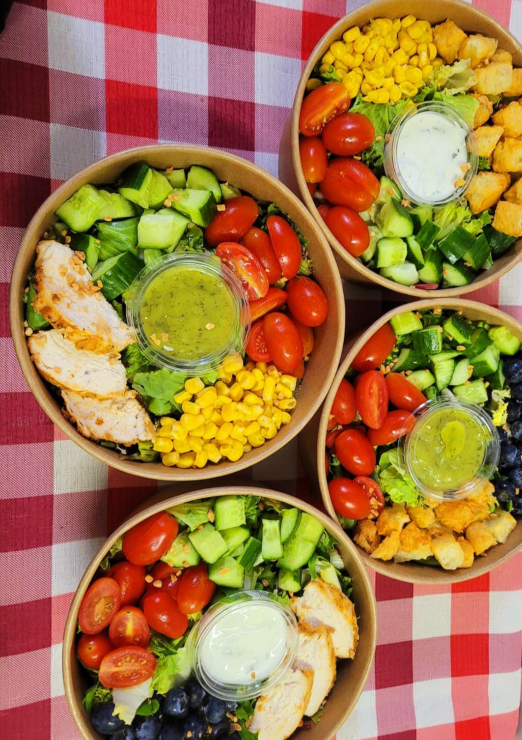 Cztery papierowe miski z różnorodnymi sałatkami: jedna z grillowanym kurczakiem, kukurydzą i sosem ziołowym, druga z grzankami i sosem śmietanowym, a dwie pozostałe z borówkami i sosem ziołowym,...