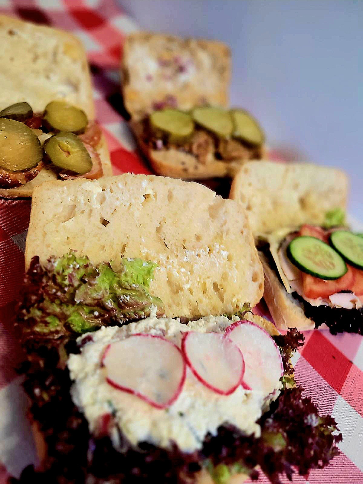 Różnorodne kanapki na jasnym pieczywie, ułożone na czerwono-białej kraciastej ściereczce, z widocznymi składnikami: ogórki kiszone, rzodkiewki, sałata, pomidory, ser i wędlina.