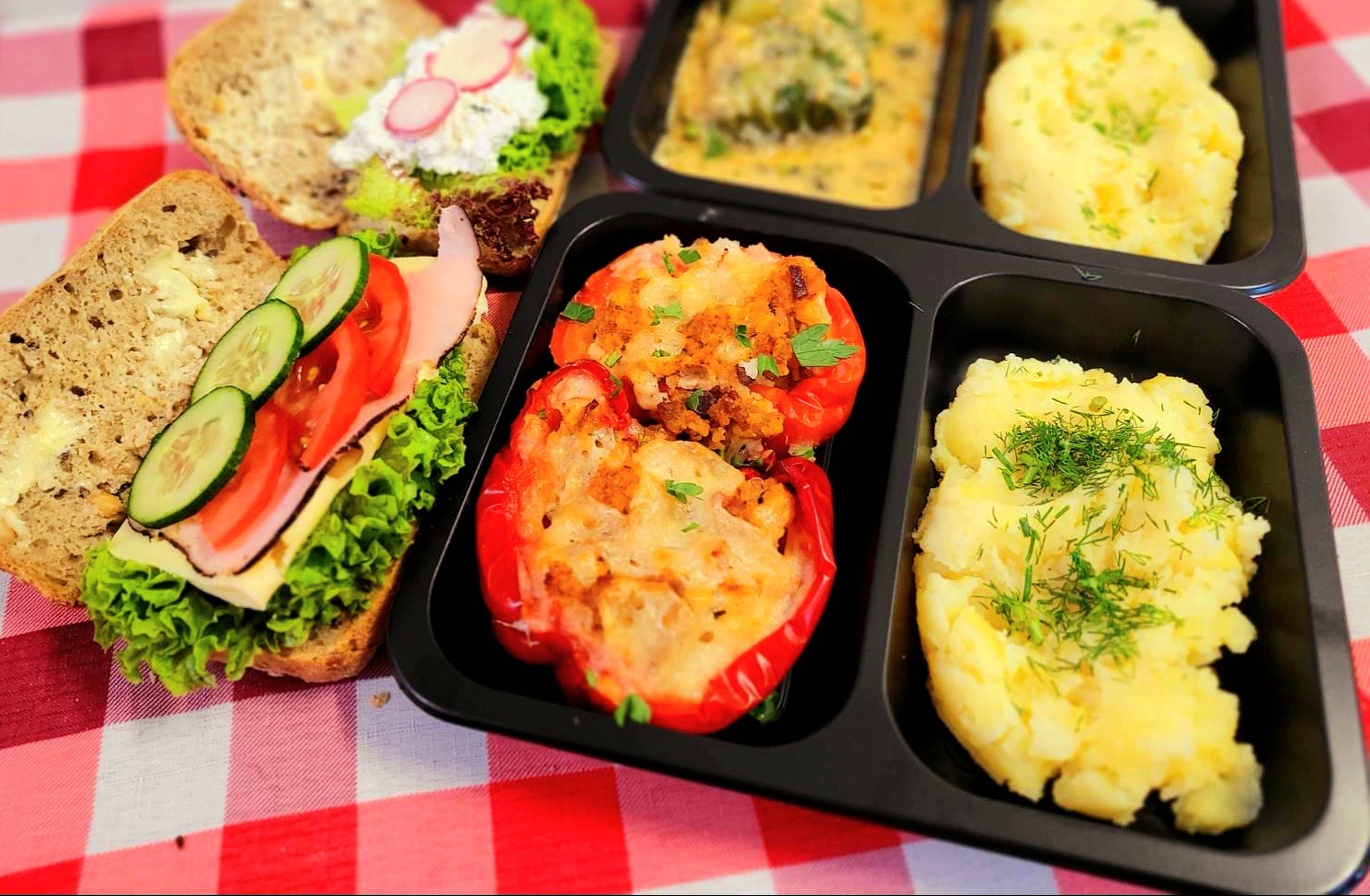 Dwa otwarte kanapki z warzywami i wędliną obok czarnego pojemnika z papryką faszerowaną i puree ziemniaczanym posypanym koperkiem, na czerwono-białej kraciastej ściereczce.