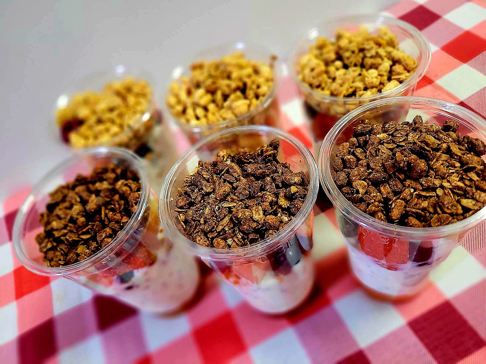 Sześć przezroczystych kubków z warstwami jogurtu, owoców i chrupiącej granoli, dwa rodzaje granoli: jasna i czekoladowa, na czerwono-białej kraciastej ściereczce.