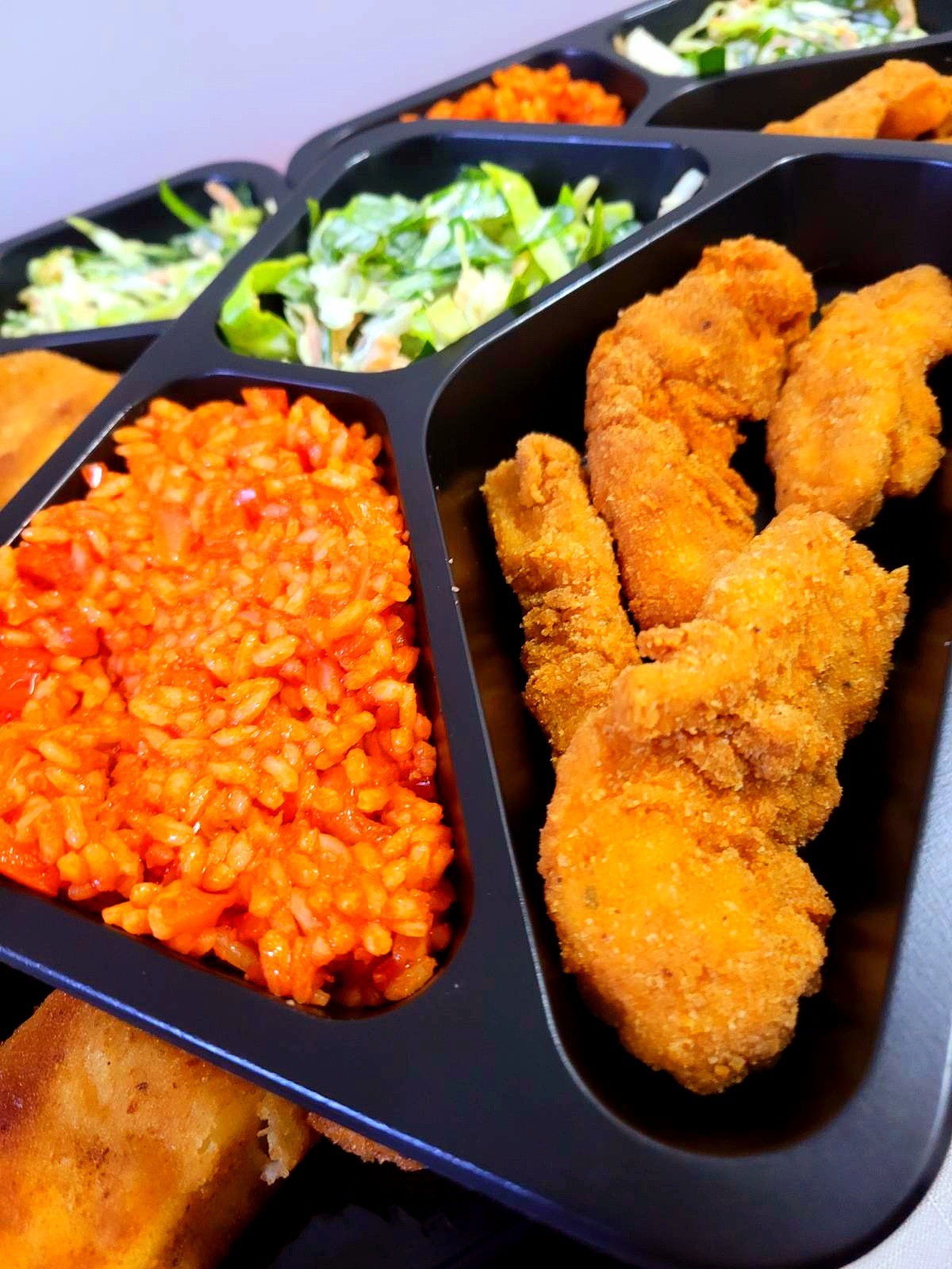 Catering dietetyczny: porcja ryżu z warzywami, panierowane kawałki kurczaka i sałatki w czarnym, podzielonym pojemniku.