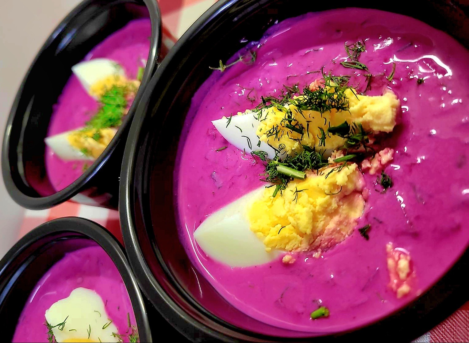 Chłodnik litewski w czarnych miseczkach z jajkiem na tarczy i posiekanym koperkiem, ustawiony na czerwono-białej kraciastej ściereczce, widok z góry.