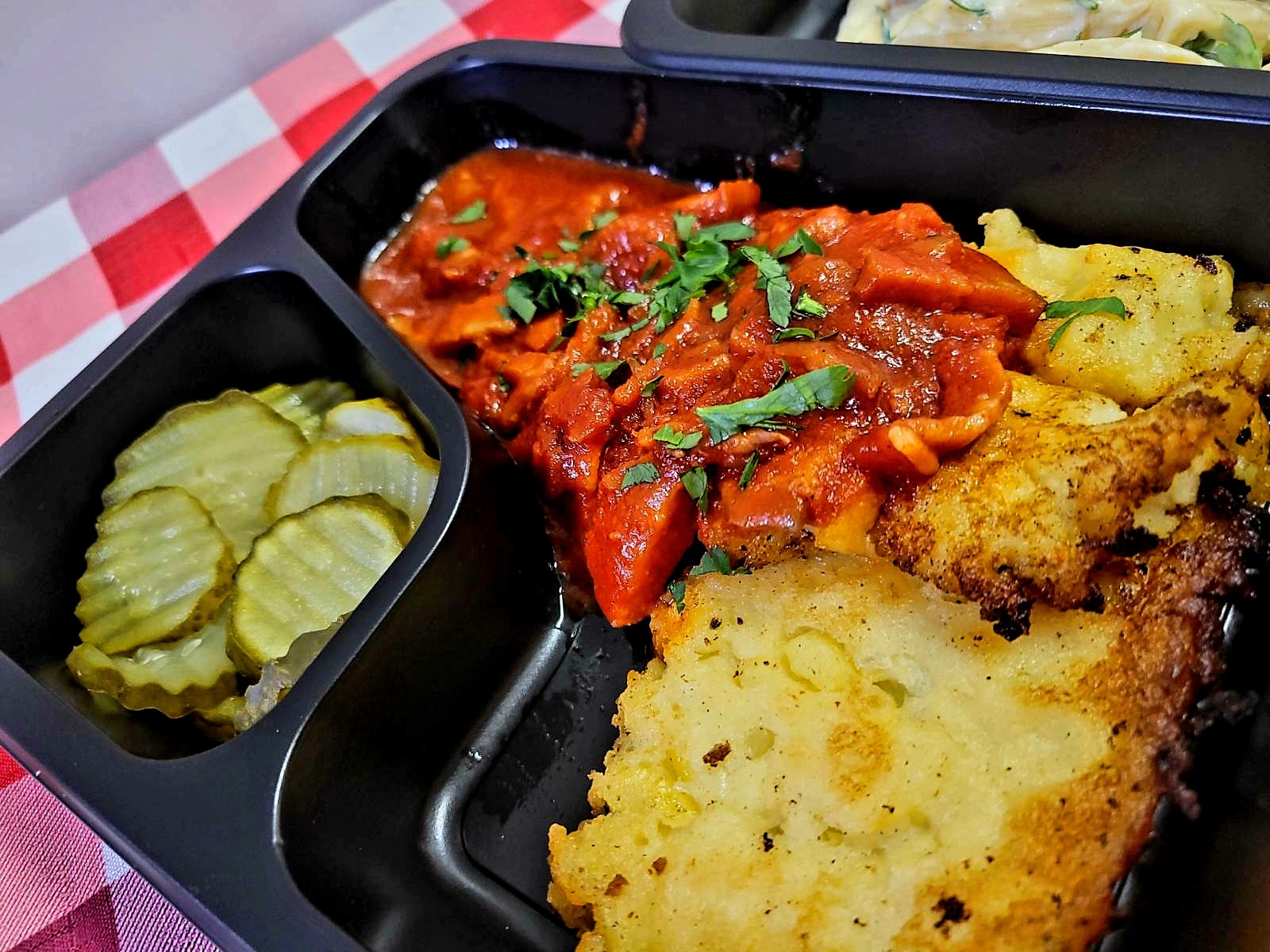 Danie obiadowe w czarnym pojemniku z trzema przegródkami: gulasz warzywny z ziołami, placki ziemniaczane i ogórki konserwowe na czerwono-białej kraciastej ściereczce.