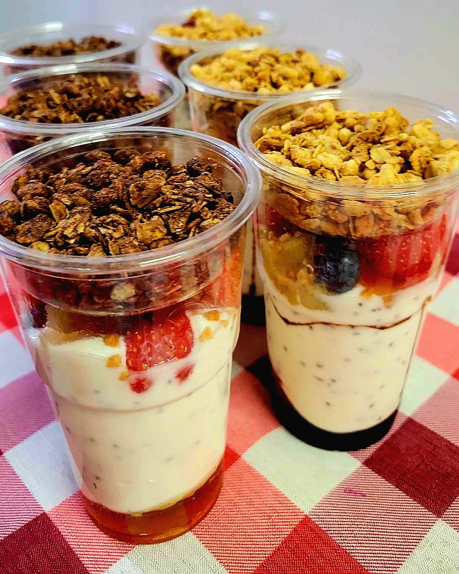Deser w przezroczystych kubkach z warstwami jogurtu chia, owoców (truskawki, borówki), miodu na dnie i posypki z granoli na wierzchu, ułożony na czerwono-białej kraciastej ściereczce.
