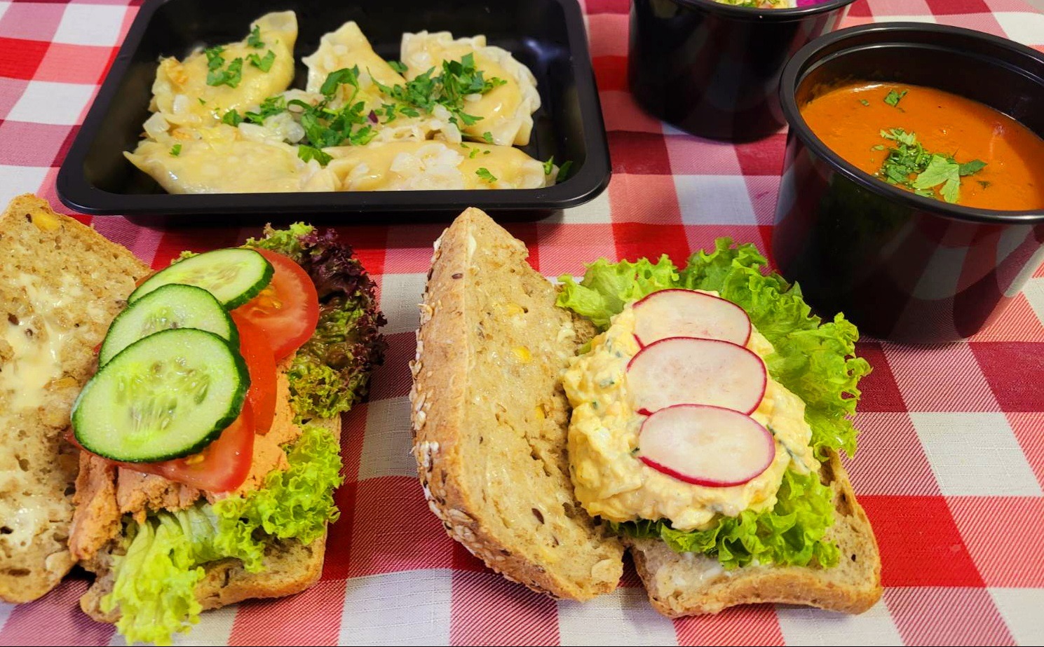 Dwa otwarte, wieloziarniste kanapki z sałatką jajeczną i rzodkiewką oraz z pastą, ogórkiem i pomidorem, pierogi okraszone cebulą i natką pietruszki w czarnym pojemniku oraz zupa pomidorowa...