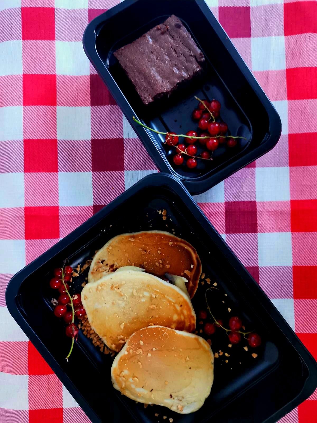 Dwa czarne pojemniki z jedzeniem na czerwono-białej kraciastej serwetce. W jednym pojemniku brownie i gałązka czerwonych porzeczek, w drugim trzy naleśniki posypane kruszonką i gałązka czerwonych...