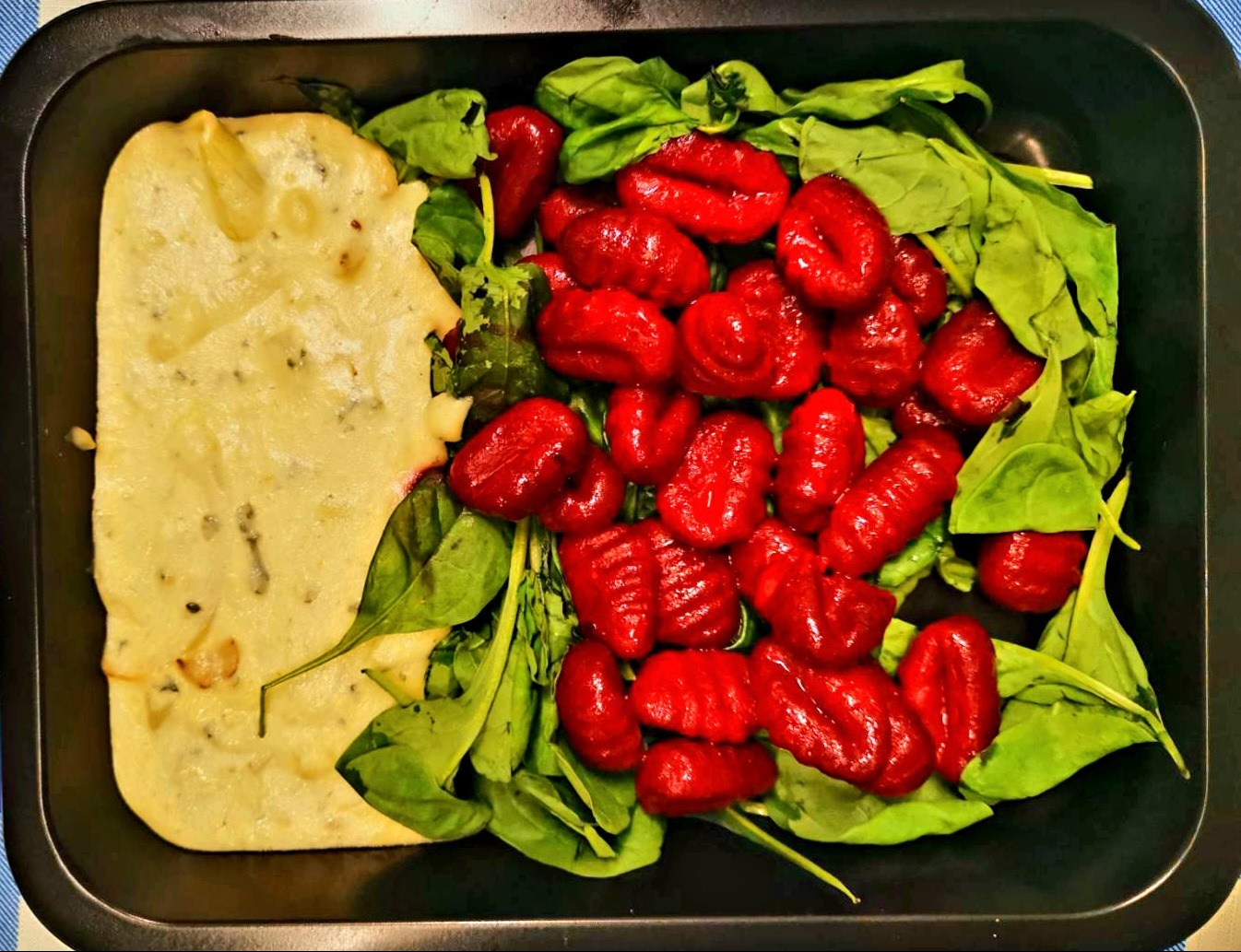 Dietetyczny posiłek w czarnym pojemniku: czerwone gnocchi na świeżym szpinaku obok zapiekanki z sosem serowym.