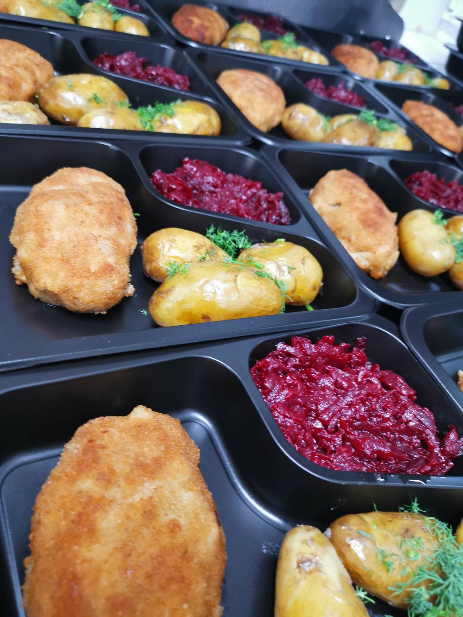 Rzędy czarnych pojemników z gotowymi daniami: kotlet panierowany, ziemniaki z koperkiem i buraczki tarte, ułożone w równych porcjach, catering dietetyczny.