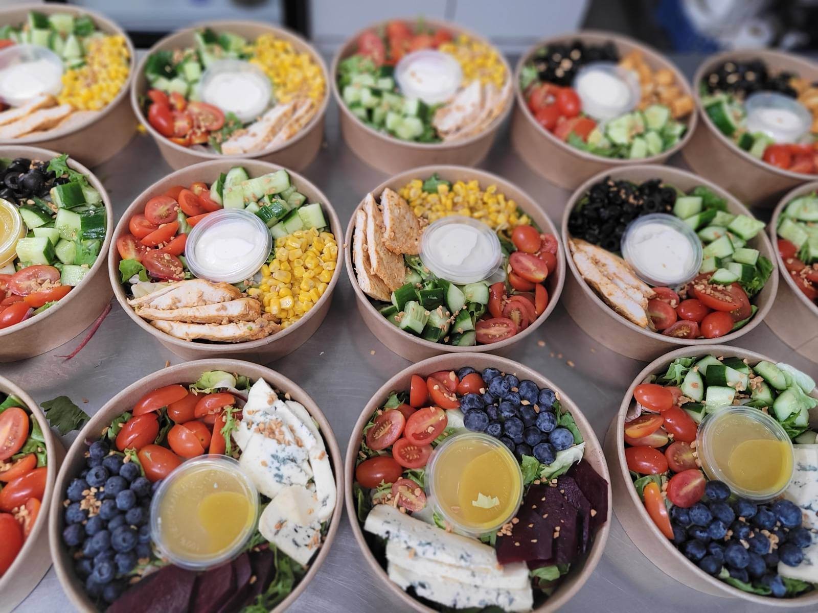 Różnorodne sałatki w papierowych miskach z widocznym kurczakiem, kukurydzą, pomidorami, ogórkami, oliwkami, serem pleśniowym, burakami i borówkami, każda z osobnym pojemniczkiem na sos.