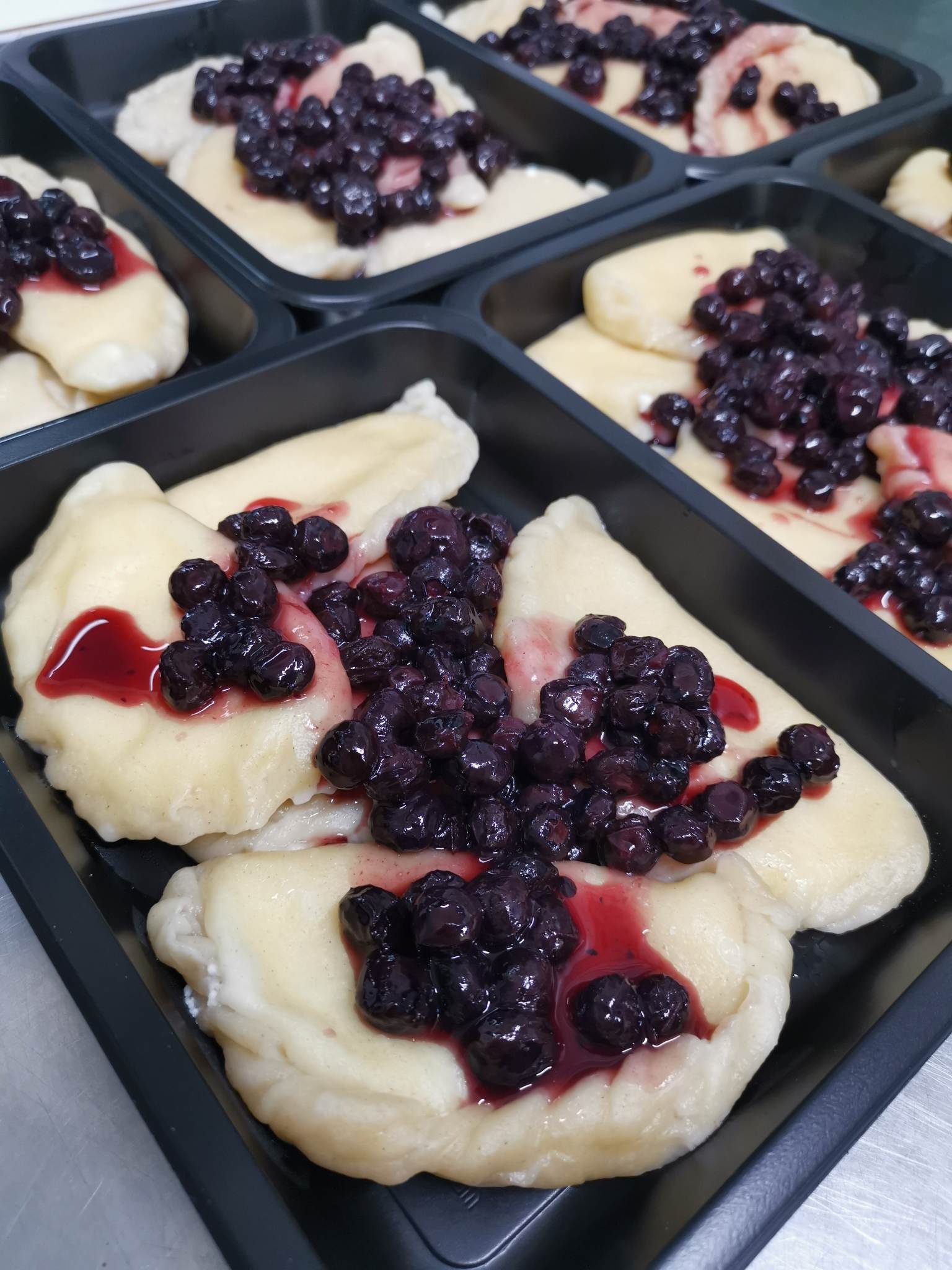 Porcje pierogów leniwych z sosem jagodowym w czarnych pojemnikach cateringowych, widoczne na metalowym blacie.